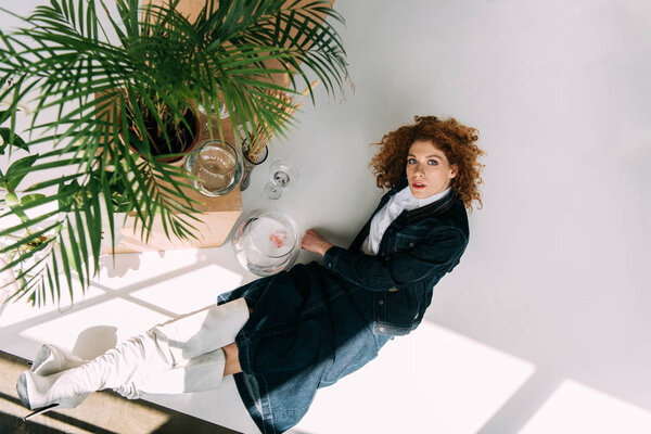 stylish redhead girl sitting and posing near wooden boxes, glasses and plants on grey with sunlight