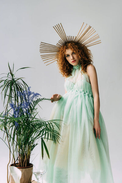 redhead girl with accessory on head in dress posing near plants on grey