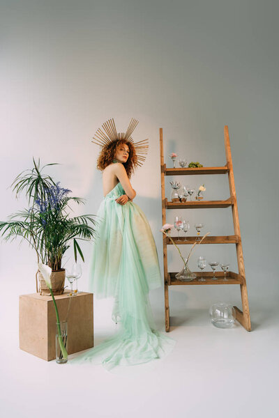 fashionable redhead girl with accessory on head posing near ladder with flowers and glasses
