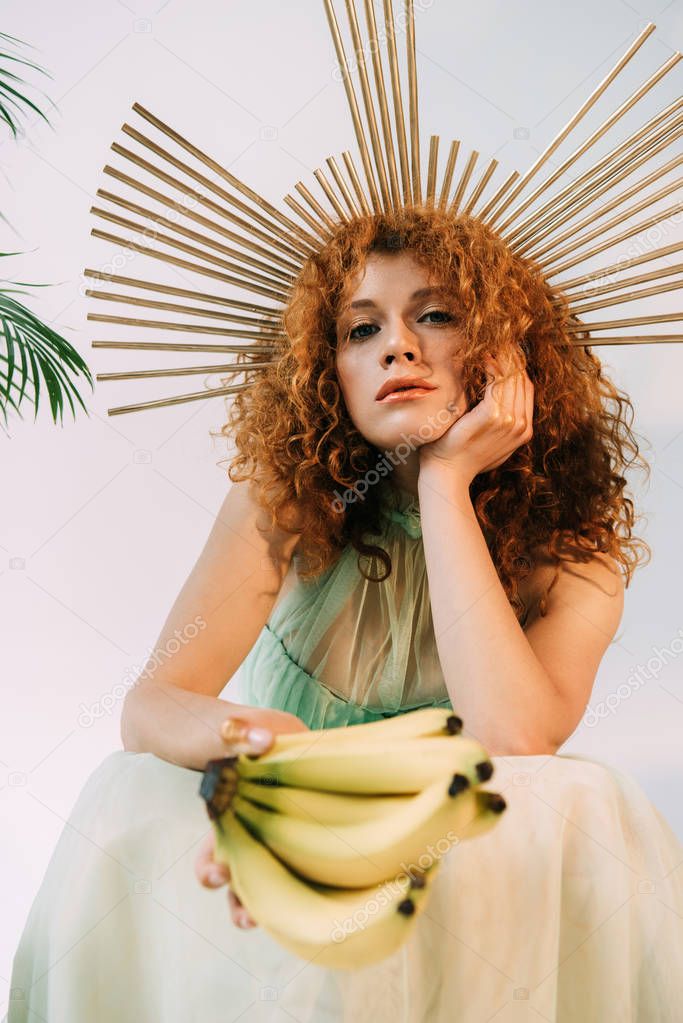 stylish redhead woman with accessory on head posing with bananas