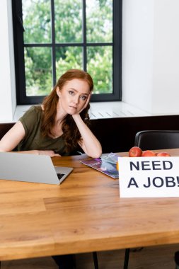 Selective focus of attractive woman looking at camera near laptop and card with need a job lettering on table  clipart
