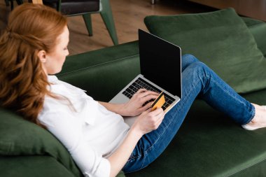 Laptop kullanan ve kredi kartı tutan kızıl saçlı kadının seçici odak noktası. 