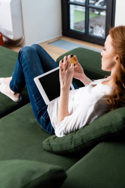 Evdeki koltukta kredi kartı ve dijital tablet tutan kadının seçici odak noktası 