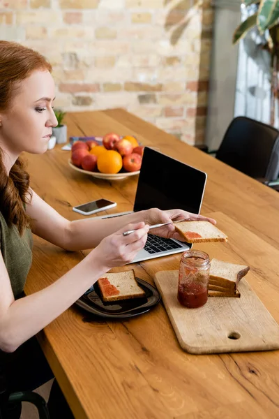 女性在餐桌上数字设备附近的吐司上倒果酱的侧视图 — 图库照片