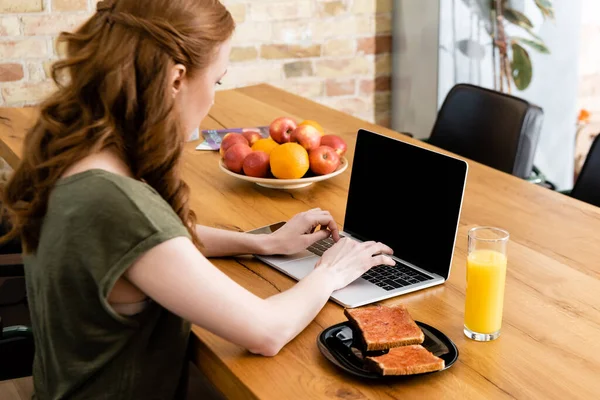 女性在烤面包附近使用笔记本电脑 桌上放上一杯橙汁的侧视图 — 图库照片