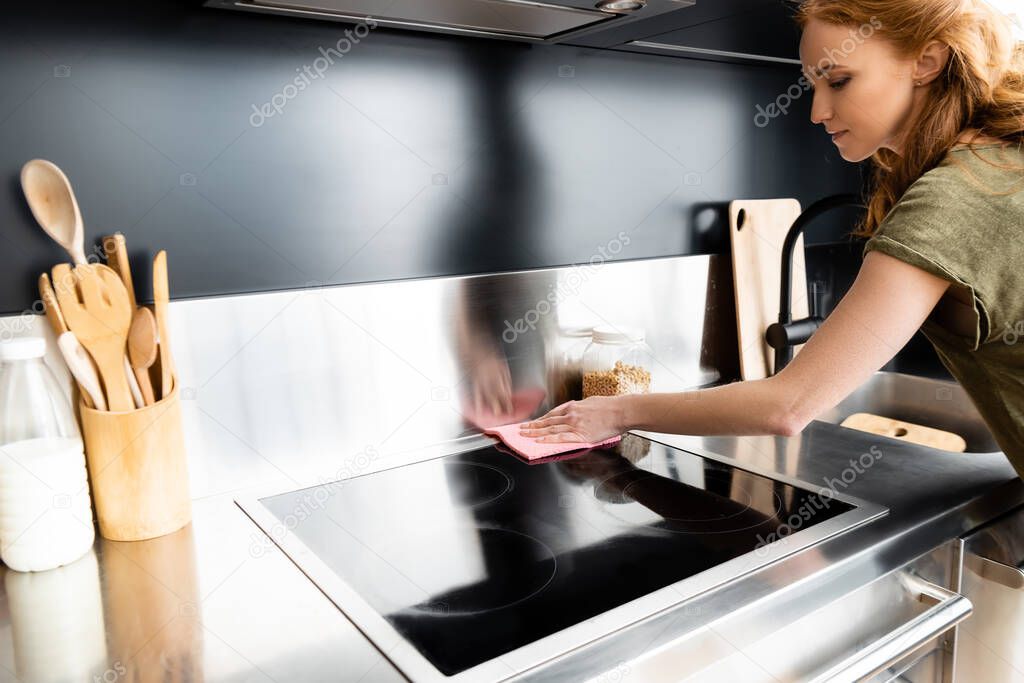 Attractive woman cleaning stove with rag in kitchen 