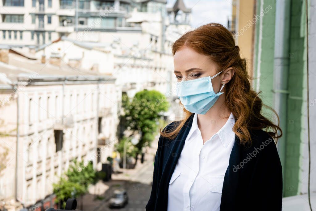 Woman in medical mask looking at urban street on terrace 