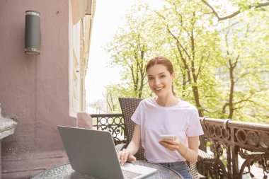 Mutlu ve çekici serbest çalışan akıllı telefon tutuyor ve balkonda dizüstü bilgisayar kullanıyor. 