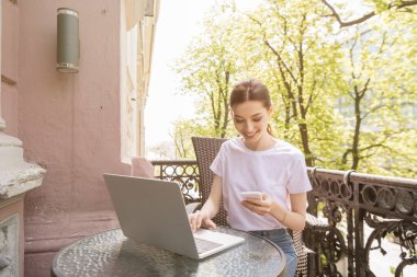 happy and attractive freelancer looking at smartphone near laptop on table clipart