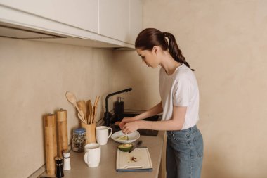 young woman putting tasty avocado in bowl near chopping board and knife  clipart