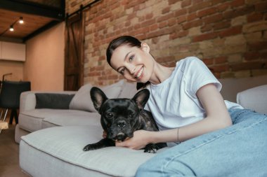 Genç bir kadın gülümsüyor ve kameraya bakıyor. Siyah Fransız bulldog 'un yanında.