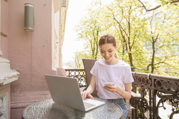 happy and attractive freelancer looking at smartphone near laptop on table