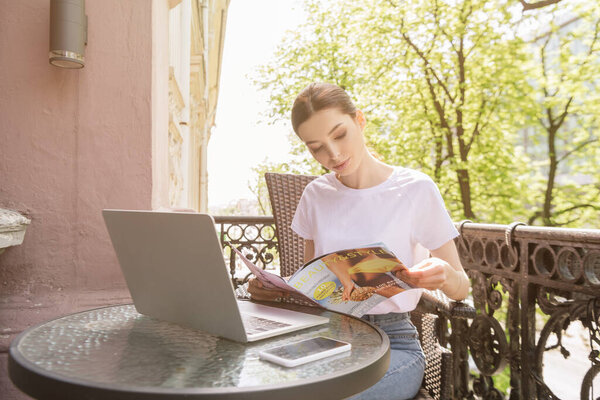 pretty freelancer reading magazine near laptop and smartphone with blank screen on table
