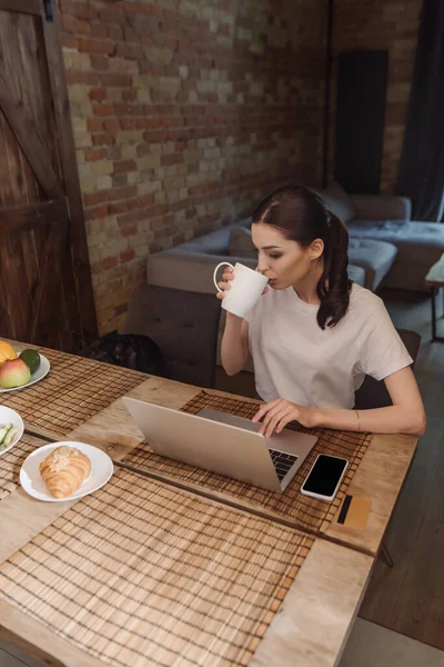 ノートパソコンの近くの魅力的なフリーランサーコーヒー空白の画面とテーブルの上のクロワッサンを持つスマートフォン — ストック写真