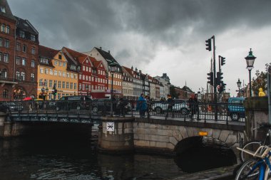 COPENHAGEN, DENMARK - 30 Nisan 2020: Nyhavn kentsel caddesi ve arka planda bulutlu gökyüzü ile kanal yakınlarındaki köprüde yürüyen insanlar  