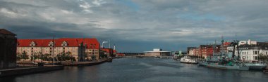 Horizontal image of buildings near canal and boats in harbor with cloudy sky at background in Copenhagen, Denmark  clipart