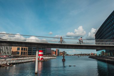 COPENHAGEN, DENMARK - 30 Nisan 2020: Nehrin üzerinde binalar ve arka planda bulutlu gökyüzü olan köprüde bisiklete binen insanlar  