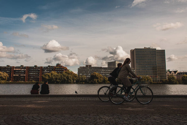 COPENHAGEN, DENMARK - APRIL 30, 2020: People riding bicycles on promenade near canal with buildings and cloudy sky at background 