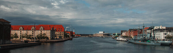 Horizontal image of buildings near canal and boats in harbor with cloudy sky at background in Copenhagen, Denmark 