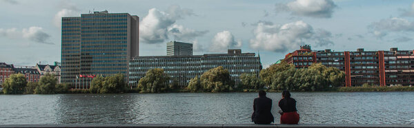 Panoramic crop of people sitting on promenade near canal with buildings and cloudy sky at background, Copenhagen, Denmark 