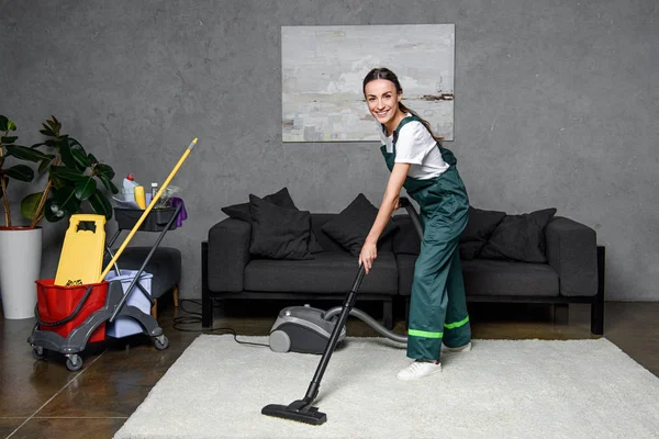 Feliz joven trabajadora de la empresa de limpieza utilizando aspiradora y sonriendo a la cámara - foto de stock