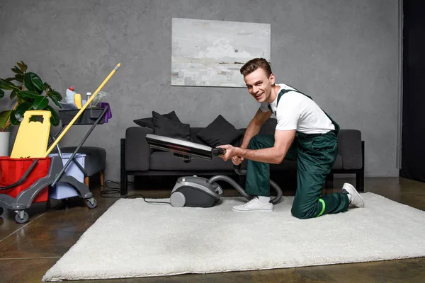 Jovem bonito usando aspirador de pó e sorrindo para a câmera, limpeza tapete profissional — Fotografia de Stock