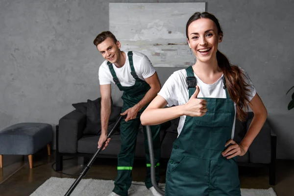 Felices trabajadores jóvenes de la empresa de limpieza utilizando aspiradora y mostrando el pulgar hacia arriba - foto de stock