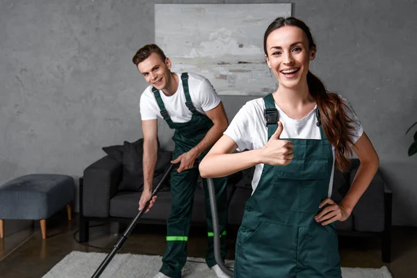 Heureux jeunes travailleurs de l'entreprise de nettoyage en utilisant l'aspirateur et montrant pouce vers le haut — Photo de stock