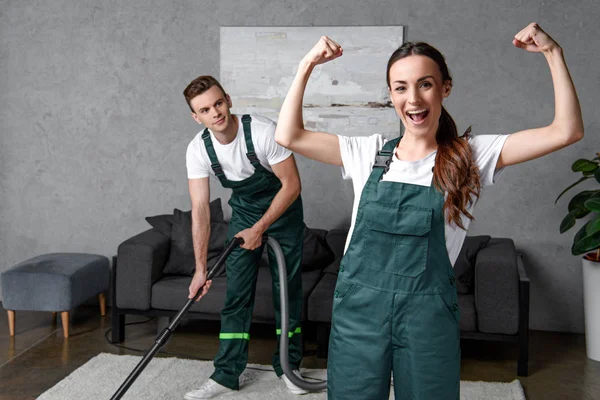 Young male cleaner using vacuum cleaner and looking at female coworker showing muscles and smiling at camera — Stock Photo