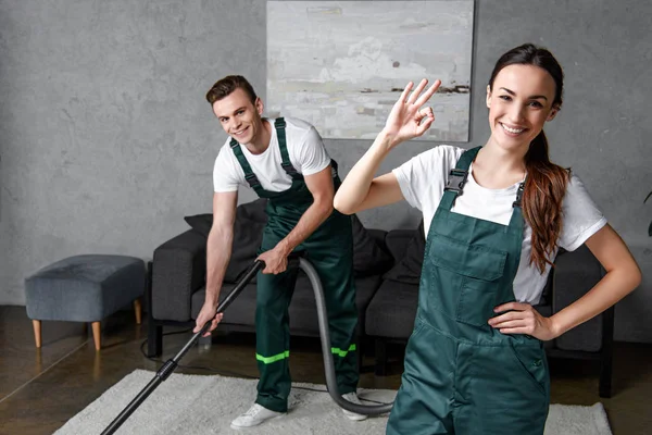 Trabajadores jóvenes felices de la empresa de limpieza utilizando aspiradora y mostrando signo ok - foto de stock