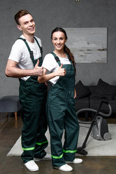 Feliz joven profesional de limpieza trabajadores de la empresa mostrando los pulgares hacia arriba y sonriendo a la cámara - foto de stock