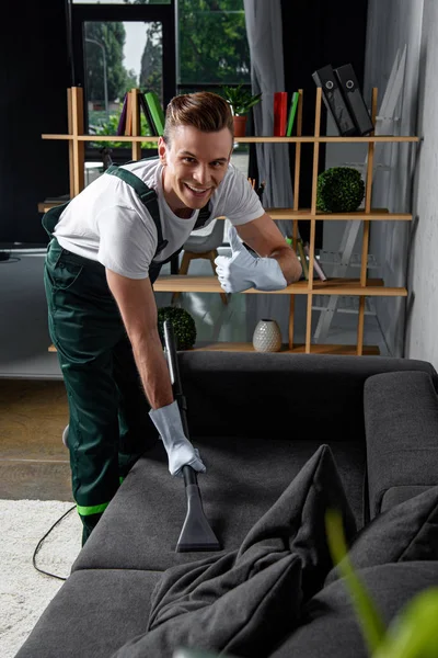 Sonriente joven empresa de limpieza trabajador limpieza sofá con aspiradora y mostrando el pulgar hacia arriba - foto de stock