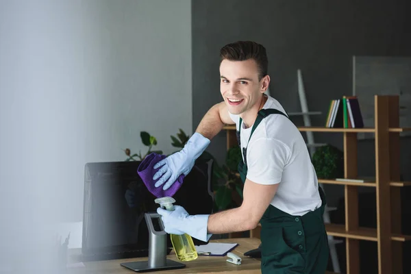 Hübsche junge professionelle Reinigungskraft lächelt in die Kamera, während sie den Computermonitor im Büro reinigt — Stockfoto