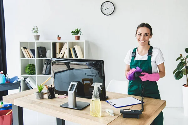 Schöne junge professionelle Reinigungskraft lächelt in die Kamera, während sie modernes Büro putzt — Stockfoto