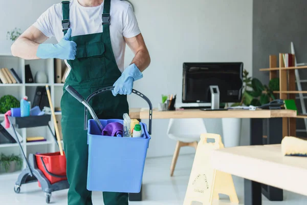 Tiro cortado de limpeza profissional segurando balde com suprimentos de limpeza e mostrando polegar para cima — Fotografia de Stock