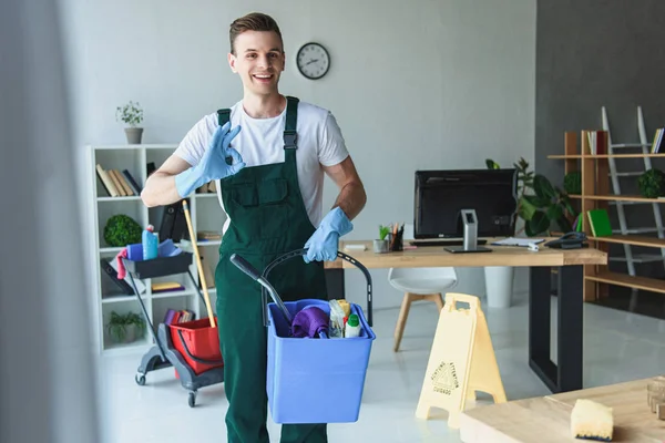 Handsome smiling young cleaner holding bucket with cleaning supplies and showing ok sign — Stock Photo