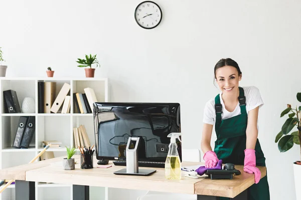 Beautiful young cleaner smiling at camera while cleaning table in modern office — Stock Photo