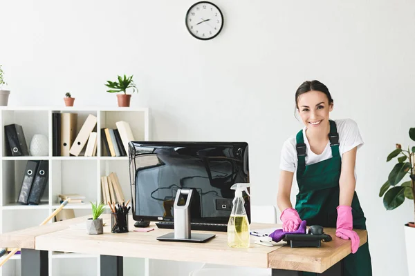 Schöne junge Putzfrau lächelt in die Kamera, während sie den Tisch im modernen Büro putzt — Stockfoto