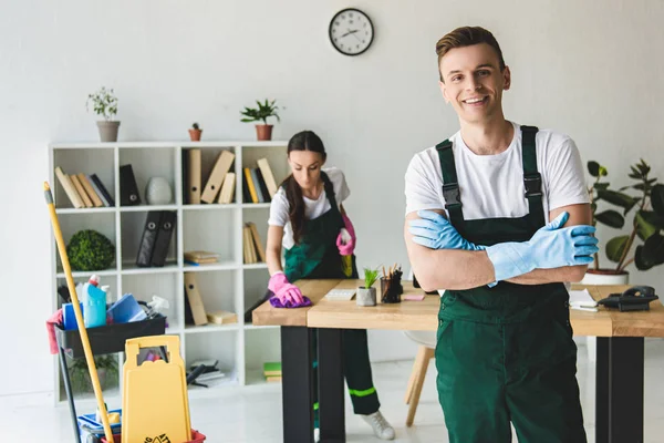 Handsome young cleaner in rubber gloves standing with crossed arms and smiling at camera — Stock Photo