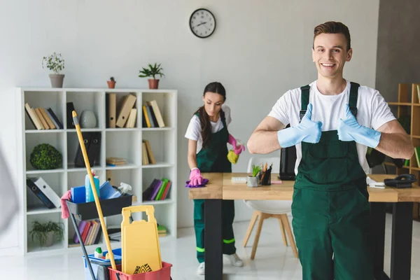 Junge männliche Putzfrau in Gummihandschuhen zeigt Daumen hoch und lächelt in die Kamera — Stockfoto