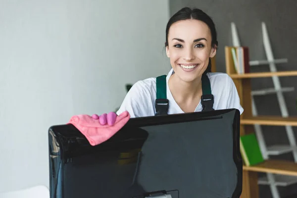 Atractiva joven limpieza monitor de ordenador y sonriendo a la cámara - foto de stock