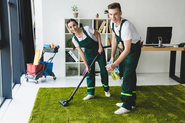 Fröhliche junge Hausmeister klappern Teppich und lächeln in die Kamera im Büro — Stockfoto