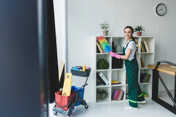 Junge Putzfrau hält Staubwedel in der Hand und lächelt in die Kamera, während sie im Büro Regale putzt — Stockfoto