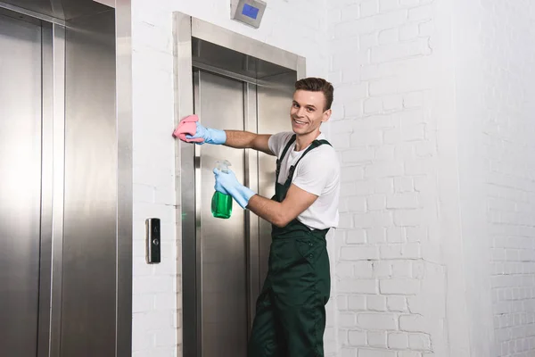 Guapo joven limpiador lavado ascensor y sonriendo a la cámara - foto de stock