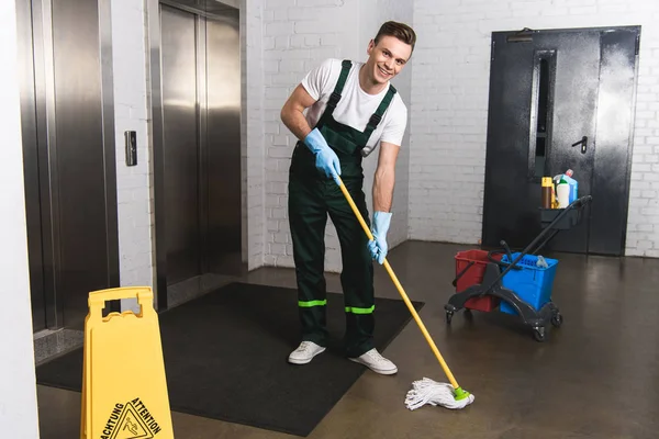 Apuesto joven conserje fregando piso y sonriendo a la cámara - foto de stock