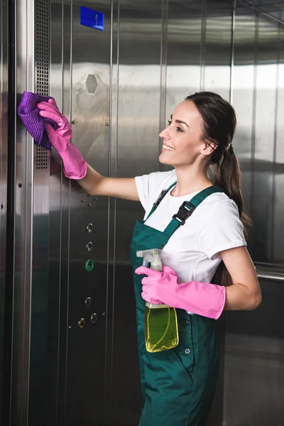 Schön lächelnde junge Reinigungskraft, die Fahrstuhl mit Spülmittel und Lappen reinigt — Stockfoto