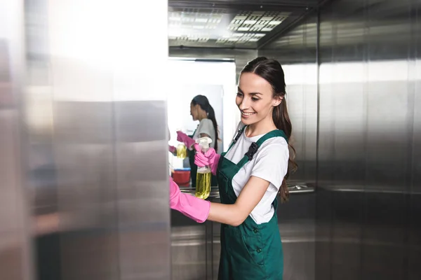 Hermoso sonriente joven conserje limpieza ascensor con detergente y trapo - foto de stock