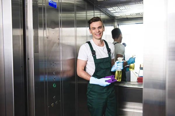 Bonito jovem zelador segurando frasco de spray com detergente e pano, sorrindo para a câmera no elevador — Fotografia de Stock