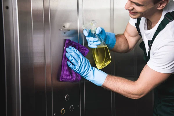 Обрезанный снимок улыбающегося молодого рабочего, чистящего лифт тряпкой и моющим средством — стоковое фото