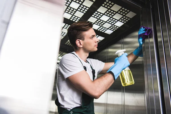 Vista de ángulo bajo del ascensor de lavado joven guapo limpiador con trapo y detergente - foto de stock